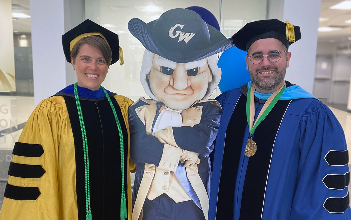 Leigh Frame and Patrick Corr dressed in graduation robes while standing next to a cardboard cutout of George